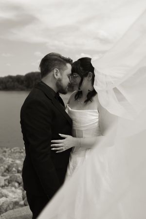 Black and white photo of bride and groom sharing intimate moment by waterfront, veil flowing in wind.