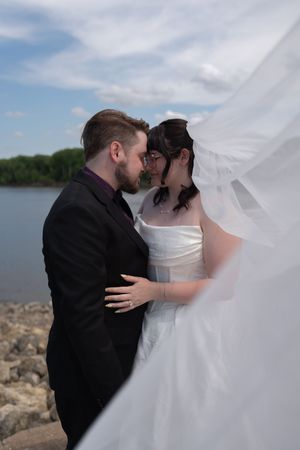 Bride and groom share romantic moment by riverside, veil billowing in wind under blue sky.