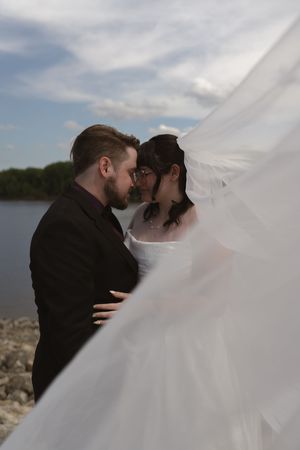 Bride and groom nearly kissing under a flowing white veil by a lakeside on their wedding day.