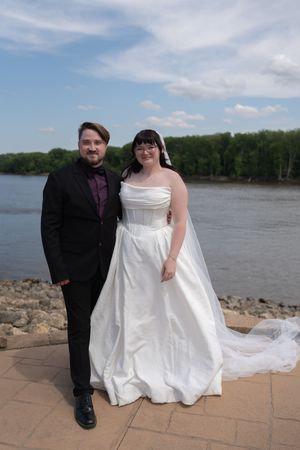 Bride and groom pose by a river on their wedding day, she in white gown, he in black suit.
