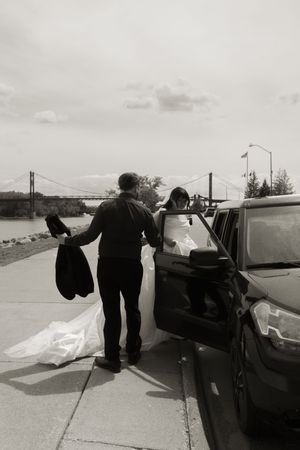 Black and white photo of groom assisting bride in wedding gown near car by waterfront.