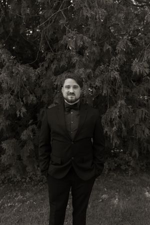 Black and white portrait of groom in black tuxedo with bow tie standing before evergreen tree.