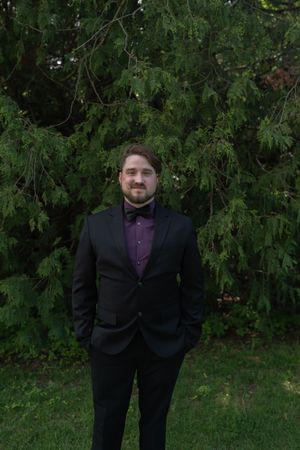 Groom in black tuxedo with purple shirt and bow tie stands before evergreen trees.