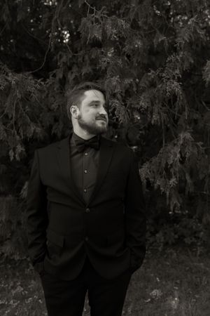Elegant black and white portrait of bearded groom in black tuxedo with bow tie standing before evergreen trees.