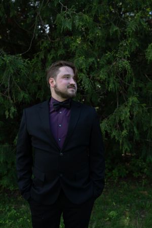 Handsome groom in black tuxedo with purple shirt and bow tie standing before lush green trees.