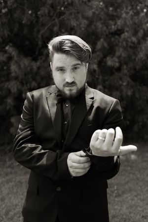 Black and white portrait of bearded groom in black suit adjusting cufflinks outdoors.