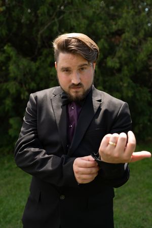 Bearded man in black suit and purple shirt adjusts cufflinks while posing outdoors at wedding.