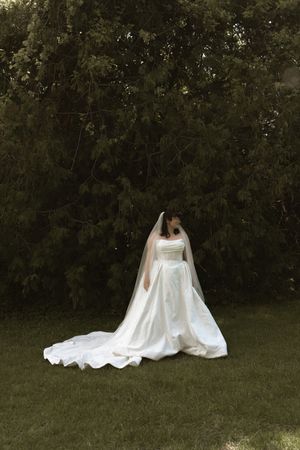 A bride in a flowing white gown and long veil stands gracefully on green grass before tall evergreen trees.