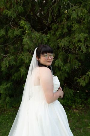Bride in white strapless gown and veil with glasses poses outdoors in front of lush green evergreen trees.