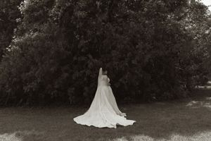 Black and white artistic photo of bride in flowing white gown and veil standing before lush trees in garden.