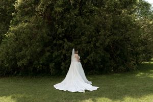 Bride in elegant white wedding gown with long train and veil standing before large green tree.