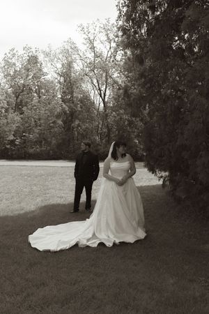 Black and white photo of bride in flowing gown and groom standing apart in lush garden setting.