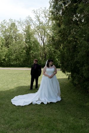 Bride in white gown with long train stands near groom in black suit in garden.