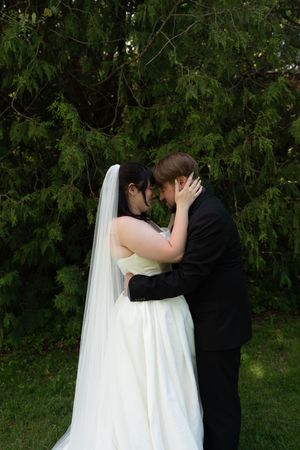Bride in white gown and veil sharing an intimate forehead touch with groom under evergreen trees.