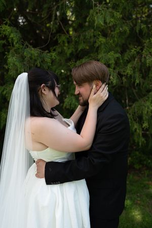 Bride and groom sharing tender embrace face to face outdoors with evergreen trees in background.
