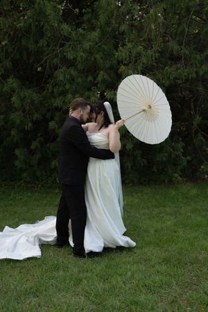 Bride and groom embrace on green lawn, she holds a white parasol, wedding train trailing behind.