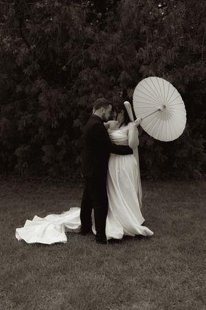 Black and white photo of bride and groom embracing with white parasol in garden.