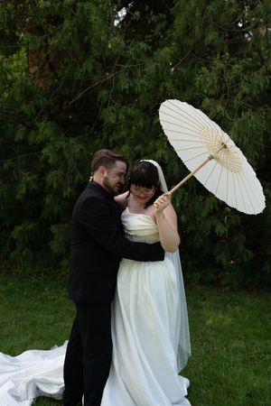 Bride and groom embrace outdoors as bride holds white parasol umbrella in green garden setting.