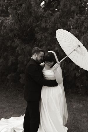Black and white photo of bride and groom embracing under a white parasol on grass.