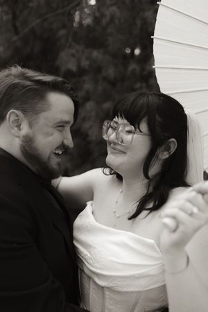 Black and white photo of laughing bride with glasses and groom sharing joyful moment with parasol.