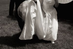 Black and white close-up of bride's flowing white wedding gown train being carried across grass.