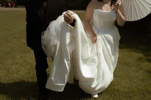 Close up of groom carrying brides white wedding dress train while walking across green lawn.