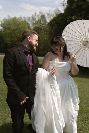 Bride and groom walk together on grass, bride holds white parasol and wedding dress train.