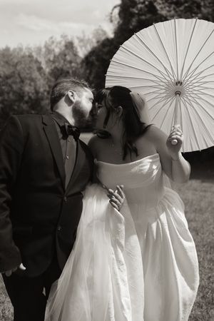 Black and white photo of bride and groom kissing while bride holds white parasol in garden.