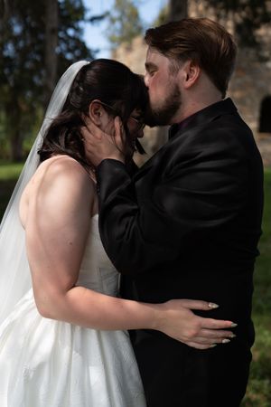 Groom in black suit tenderly kisses bride in white wedding dress and veil outdoors.