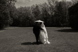 Black and white photo of bride and groom embracing with parasol in lush garden.