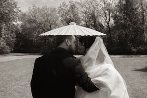 Black and white photo of bride and groom kissing under white parasol in garden setting.