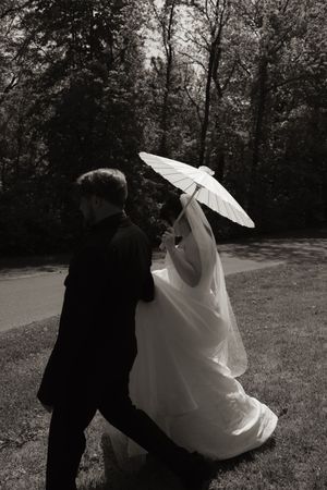 Black and white photo of bride and groom walking in a park, bride holding a white parasol and veil flowing.