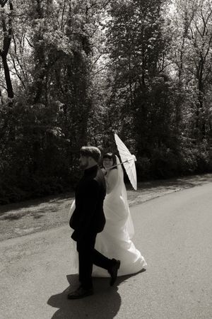 Black and white photo of bride and groom walking together on a path, bride holding white parasol.