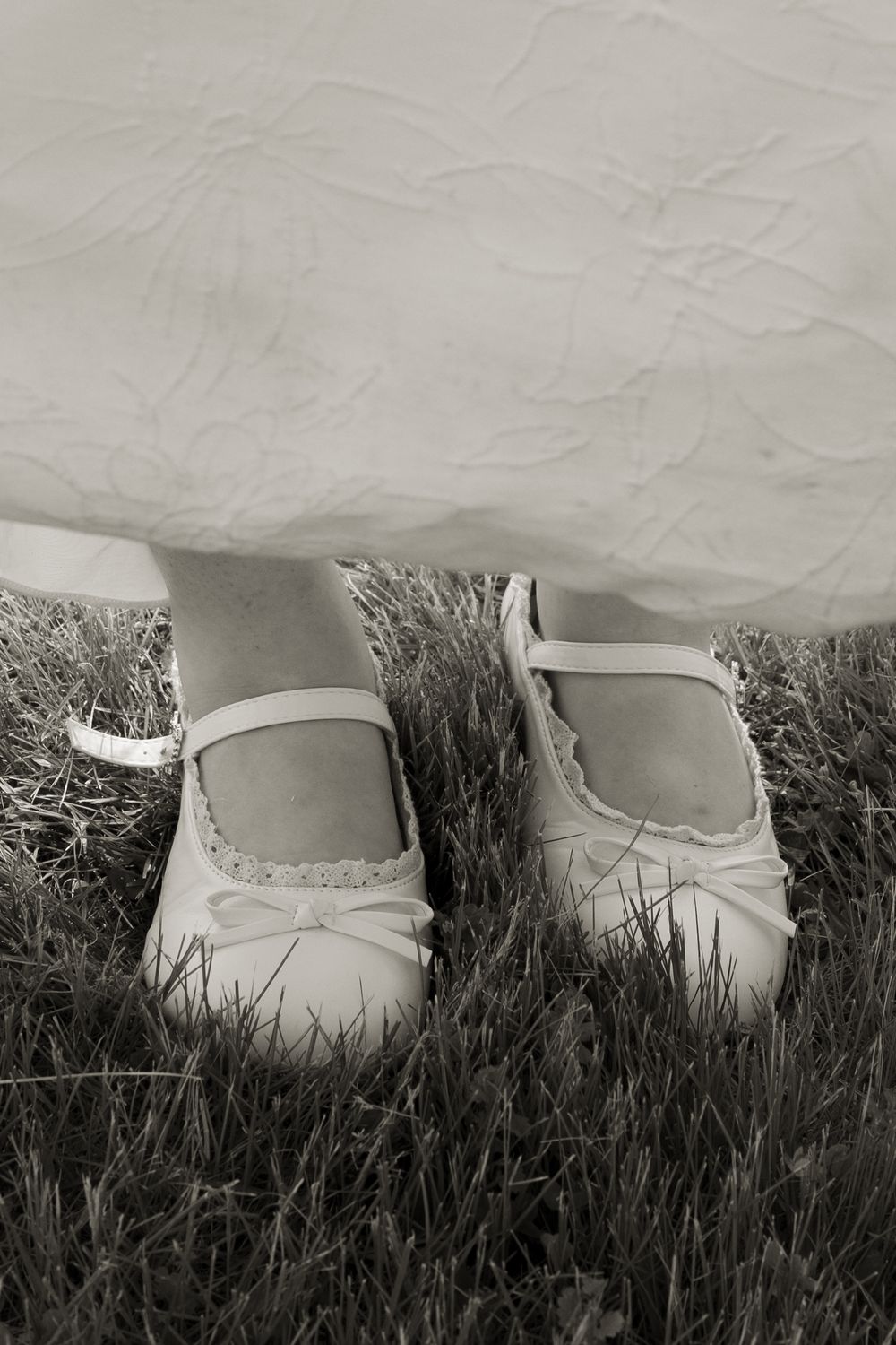 Sepia tone close-up of bride wearing white Mary Jane shoes standing on grass beneath wedding dress.