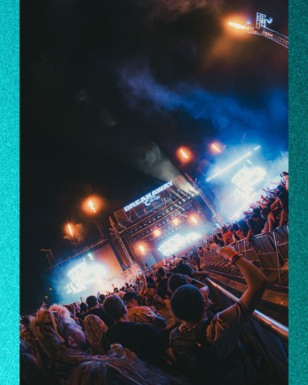 Crowd at an outdoor music festival at night with bright stage lights illuminating performers under a smoky sky.