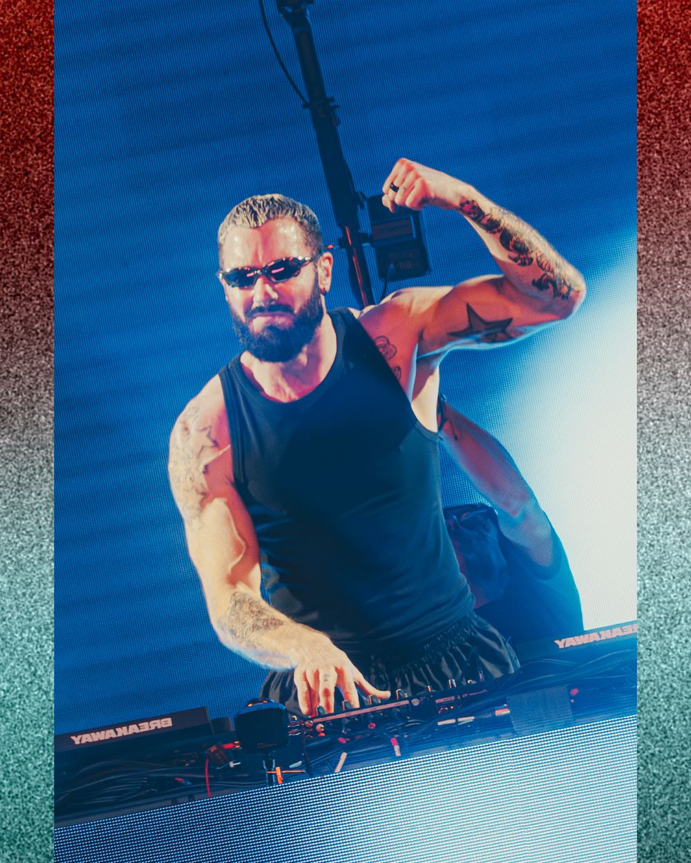 Tattooed male DJ performing on stage, wearing a black tank top, raising his fist while mixing at a DJ booth under blue stage lighting.