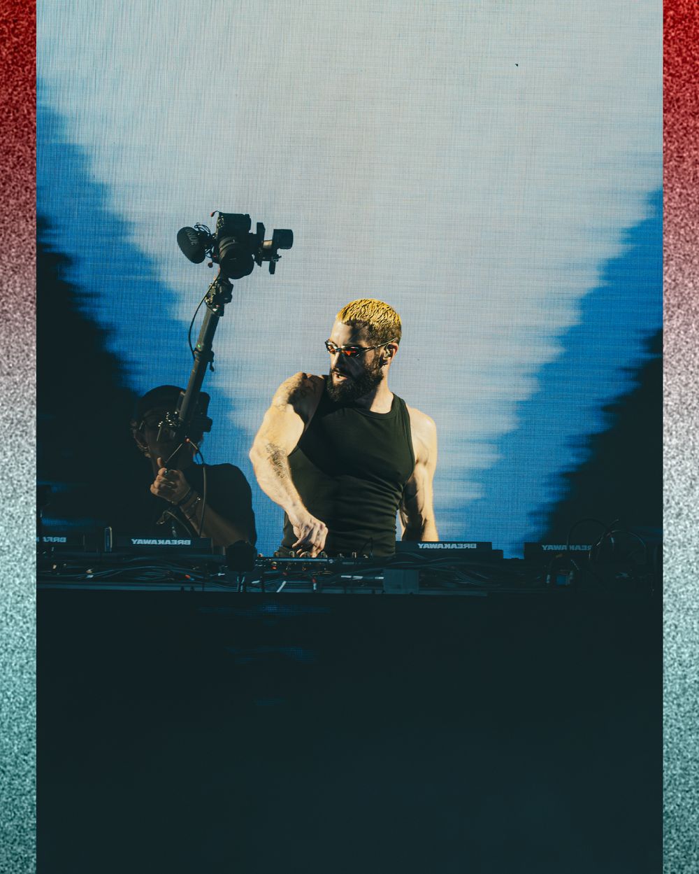 A muscular DJ performs on stage under dramatic blue lighting with a camera crane visible behind him.