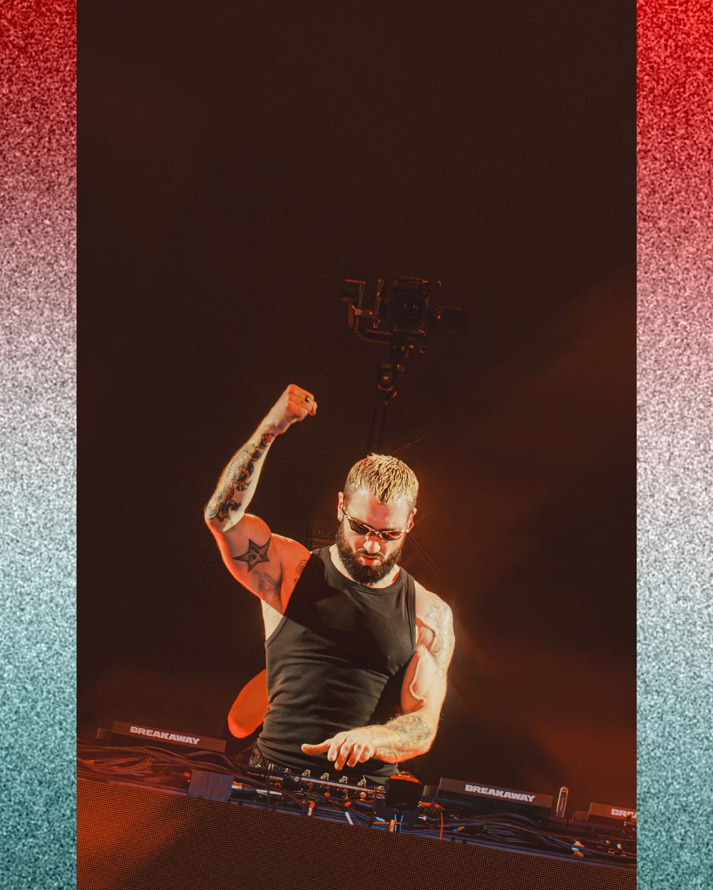 Tattooed male DJ performing on stage with fist raised, surrounded by DJ equipment under dramatic red-orange lighting.