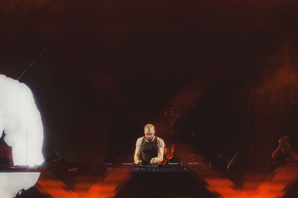 DJ performing on stage with dramatic red and orange lighting behind a mixing console.