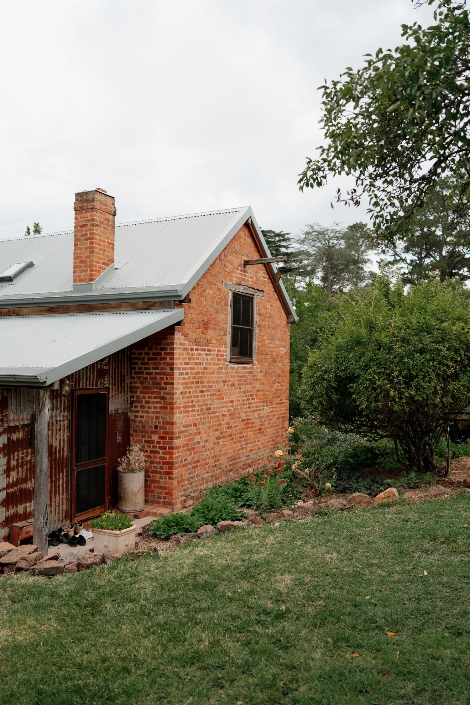 Rustic red brick cottage with metal roof, chimney, and lush garden surroundings in a rural Australian setting.