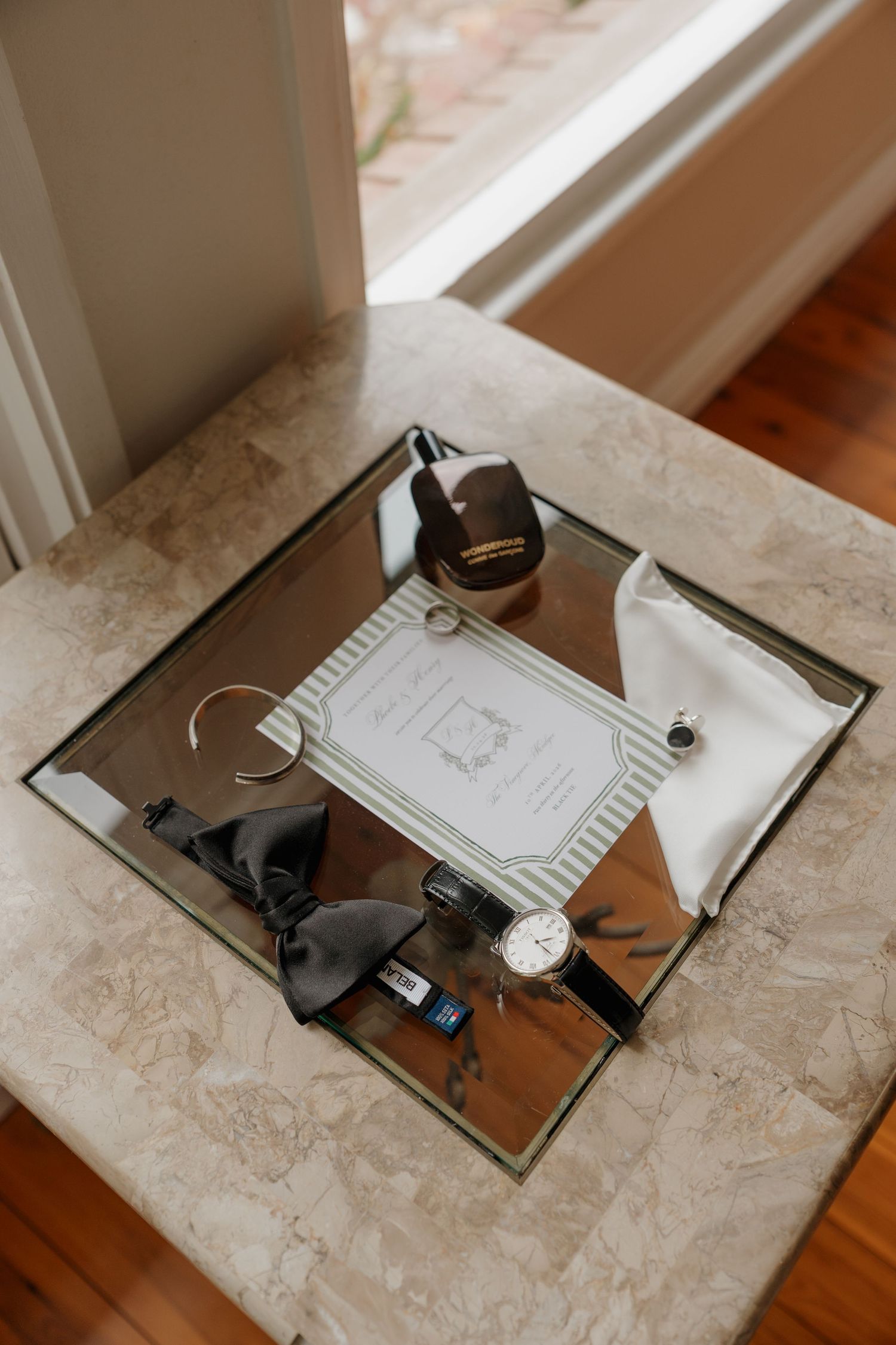 Black and white flat lay of Belance watch, bow tie, cufflinks, and invitation on marble surface.