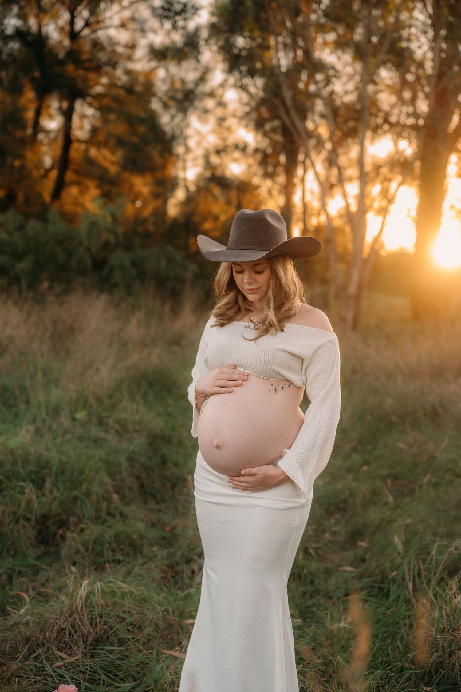 Pregnant woman in white off-shoulder gown and cowboy hat cradling baby bump at golden sunset in a field.