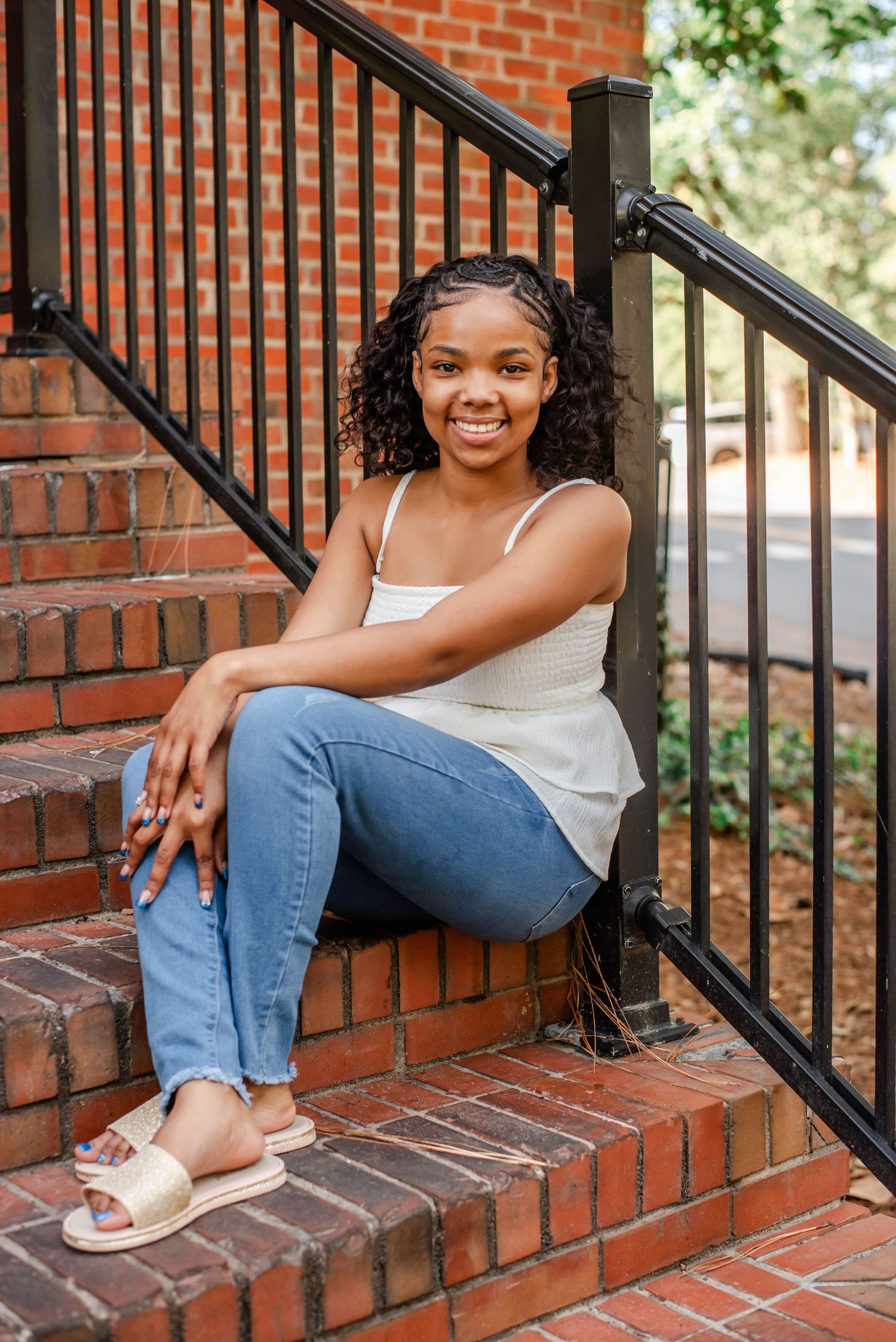Young woman smiling on brick steps, wearing white tank top, blue jeans, and gold sandals, with curly hair.