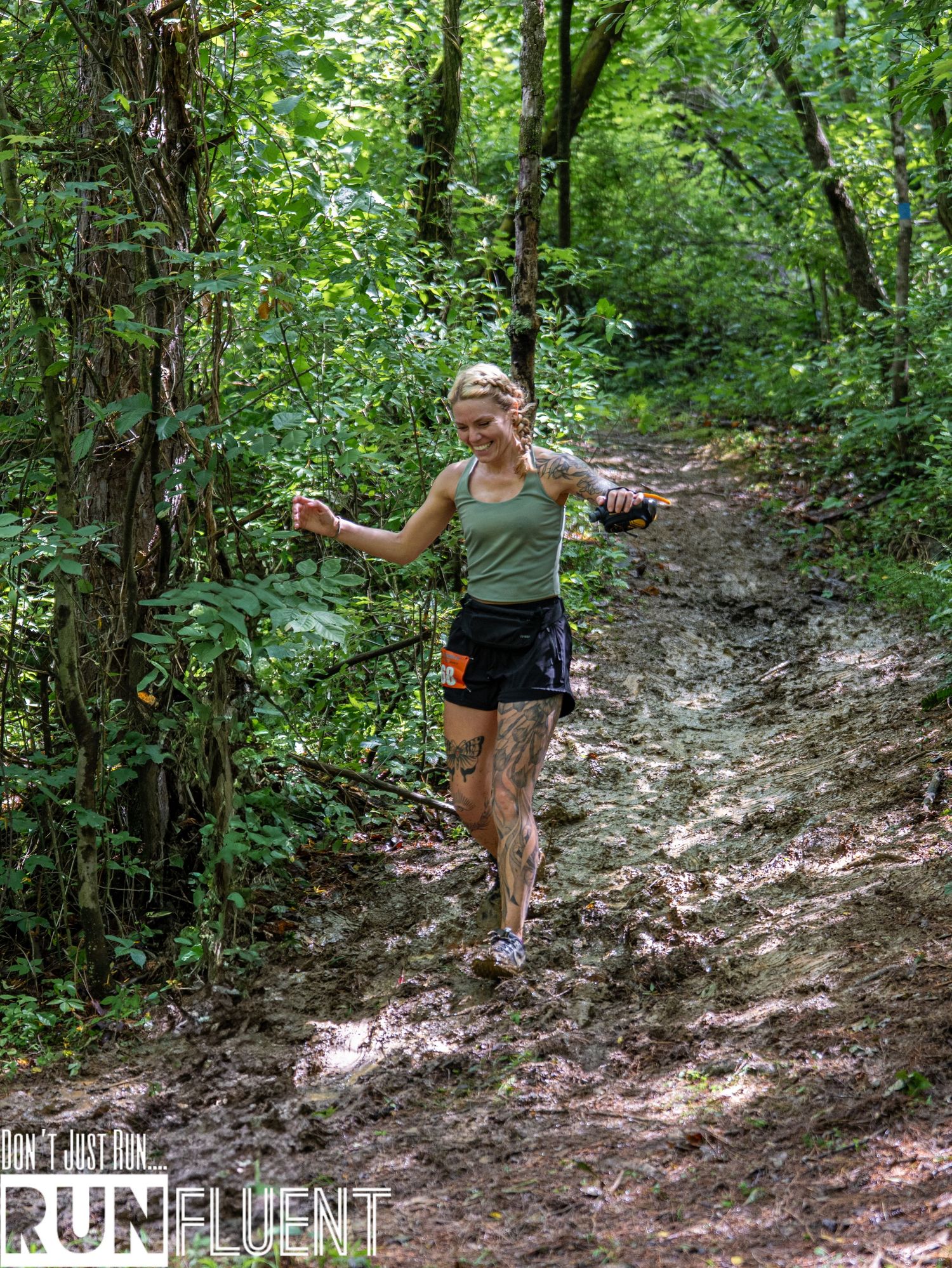 Two trail runners excitedly jump into the air on a scenic mountain path surrounded by lush greenery.