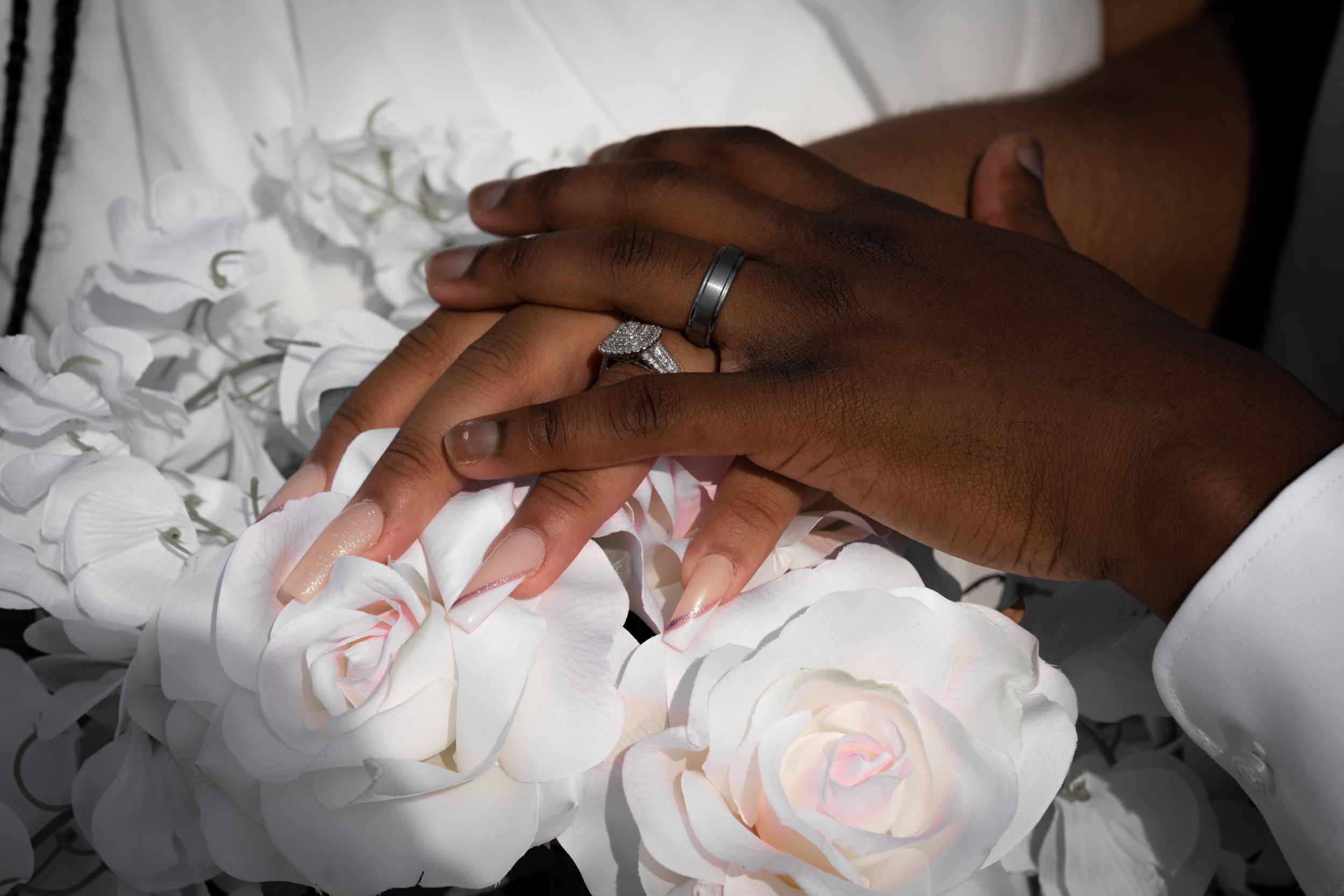 Dark and light skinned hands touch over delicate pink and white roses in an intimate wedding scene.