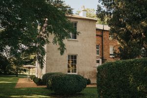 Corner view of a historic stone mansion with tall windows and manicured hedges behind leafy trees at Aswarby Rectory.