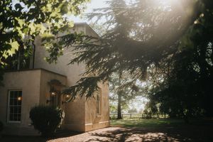 Sunlight filters through trees onto a beige stucco house exterior with windows on a tranquil afternoon at Aswarby Rectory.