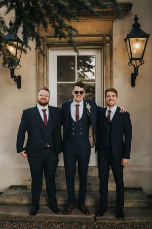 Three formally dressed men in suits stand together on elegant stone steps in front of a doorway with vintage lanterns at Aswarby Rectory.