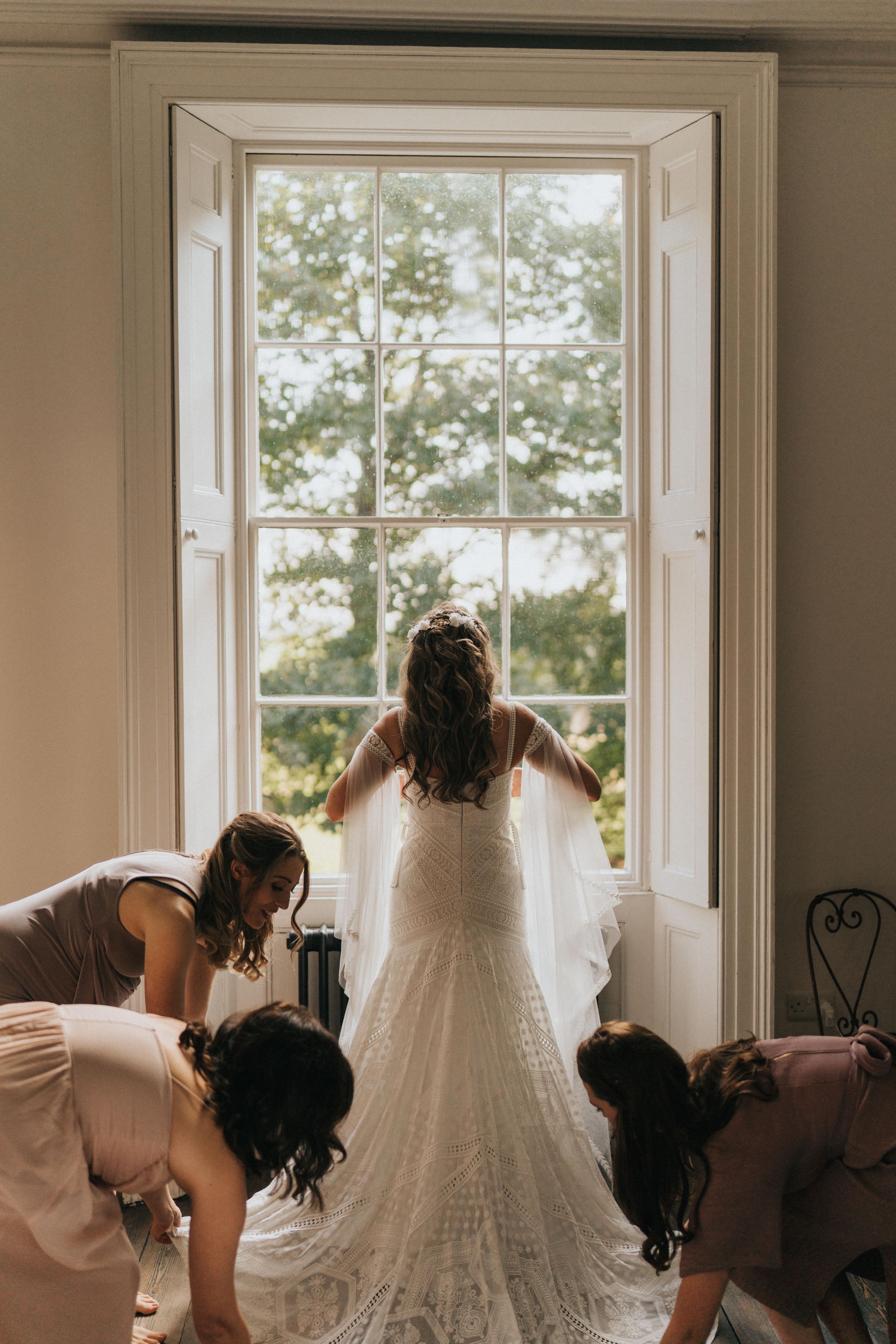 Bridesmaids help with the train of a lace wedding dress as the bride looks out a tall vintage window at Aswarby Rectory.