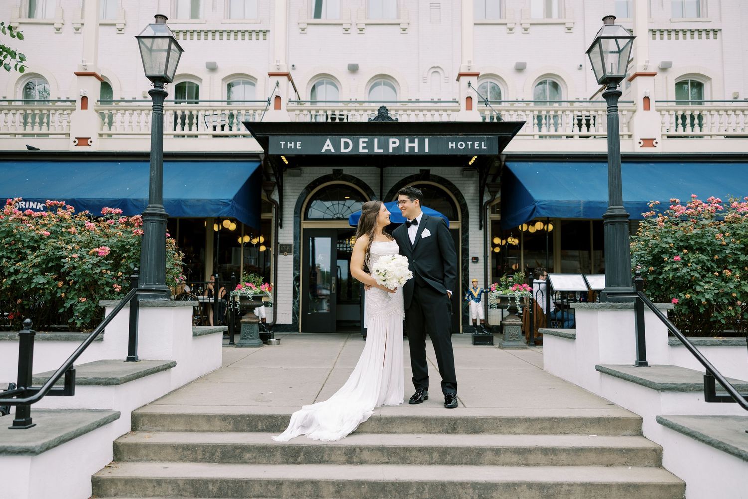 Bride and groom stand on grand stone steps of historic Adelphi Hotel in Saratoga Springs surrounded by elegant architectural details.